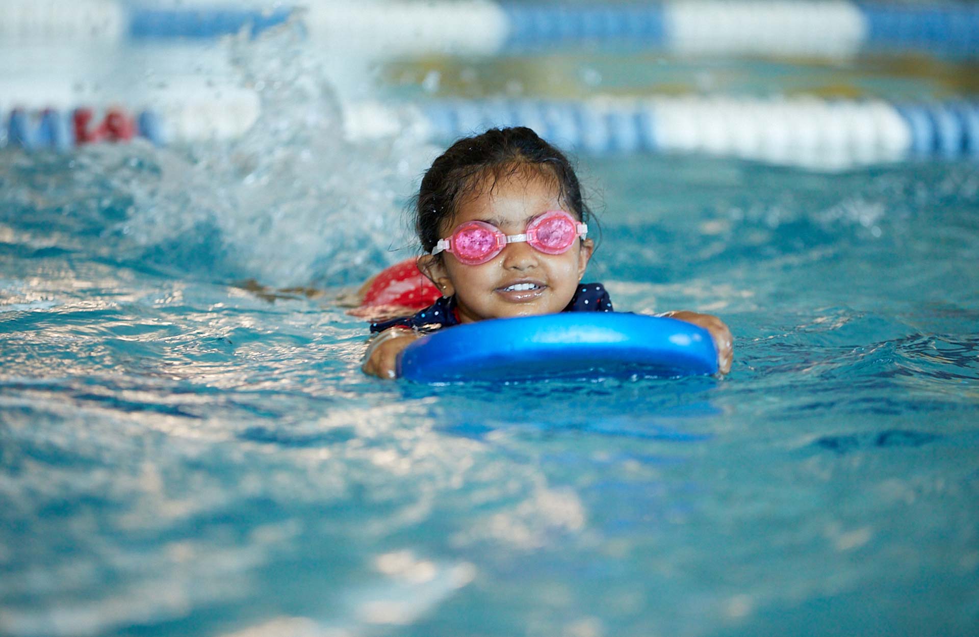 young Indian child wearing goggles using swimming kickboard in pool during swim lesson