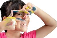 A smiling Hispanic girl looking through her paint covered hands making a box shape