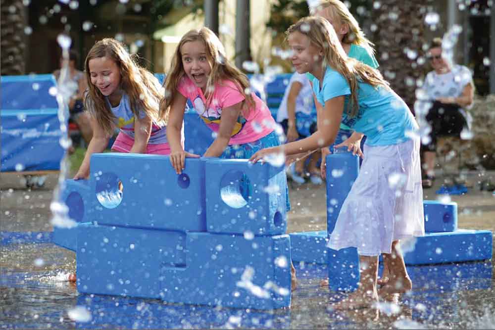 Four young girls playing with large blue building blocks in a splash pad